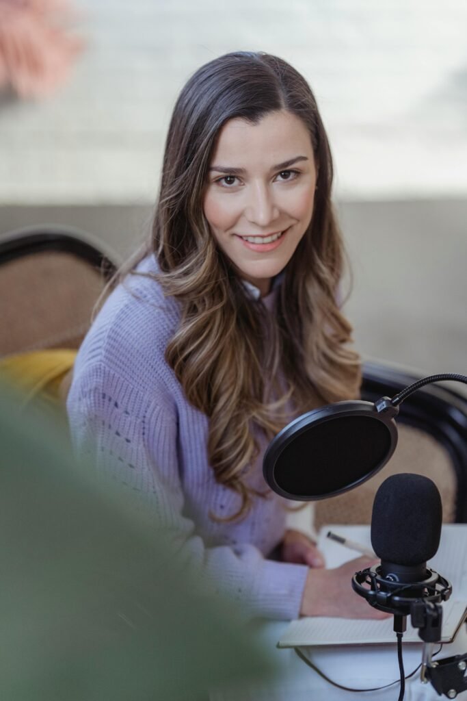 pexels-photo-6953917-6953917 A smiling young woman recording a podcast at home with microphone and notebook.